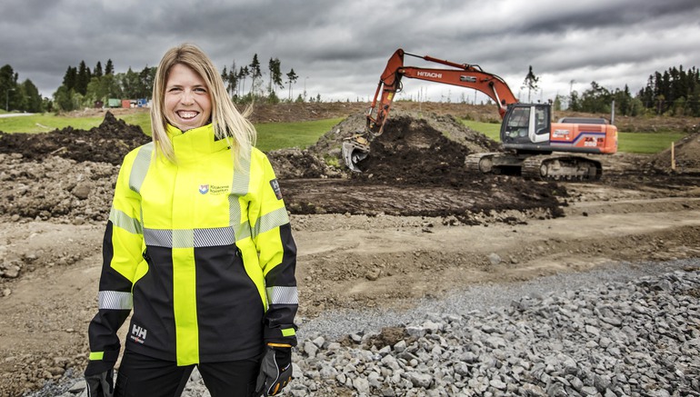 Bild på leende kvinna i byggkläder med en grävmaskin i bakgrunden