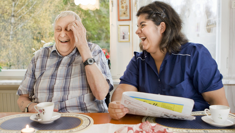 Äldre man och kvinna från hemtjänsten skrattar tillsammans vid köksbordet