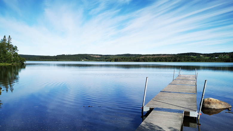 En sjö med brygga. Blankt vatten där den blåa himmlen med slöjmoln spelar sig.