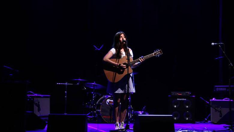 Bild på en tjej med gitarr som sjunger från en mörk scen med scenbelysning