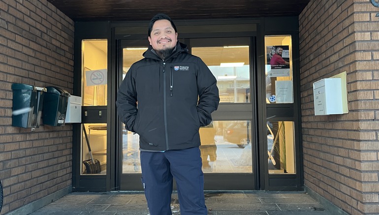 Glad man står framför ingången till en offentlig byggnad. Han är klädd i en svart vinterjacka med Krokoms logga på. 